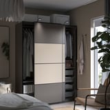 Moderne slaapkamer met PAX kledingkast in zwart/wit, schuifdeuren, hangende kleding, legplanken en een rieten stoel in de buurt.