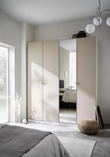Lichte, minimalistische slaapkamer met een grote beige PAX-kledingkast met spiegeldeuren, een ronde geweven kruk en een moderne kroonluchter.