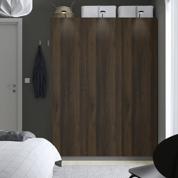 Houten PAX kledingkast, donkere afwerking, drie deuren, naast het bed, bekroond met witte dozen en hoed. Blauw item en lamp aan de muur. Hoog, verticaal brandpunt.