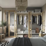 Slaapkamer met open kledingkast met verschillende kledingstukken, opbergdozen en een handtas op planken, gezien vanuit een bed met een hangende hanglamp erboven.