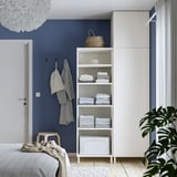 Moderne slaapkamer met witte PLATSA opbergers, handdoeken, zeegras mand, rieten handdoek en groene planten tegen blauwe muur.