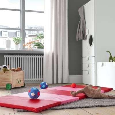 Kinderspeelkamer met rode PLUFSIG-matten, zacht speelgoed en houten opbergers. Matten zijn opgevouwen, gestapeld en kleiner dan het raam en de radiator.