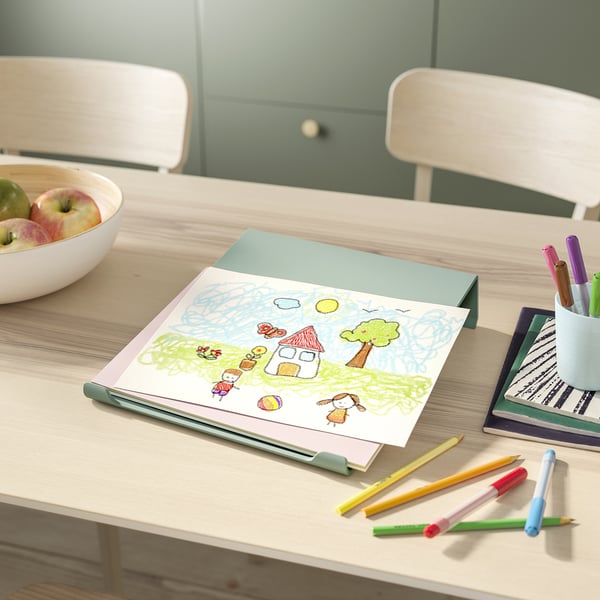 Op een lichthouten bureau zitten een kindertekening, kleurpotloden en een schaal met appels. In de buurt staan twee stapelbare stoelen.