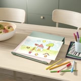 Op een lichthouten bureau zitten een kindertekening, kleurpotloden en een schaal met appels. In de buurt staan twee stapelbare stoelen.