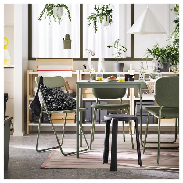 Lichte eetkamer met gebroken wit tapijt, groene stoelen, houten tafel, hangplanten en planken met decorartikelen.