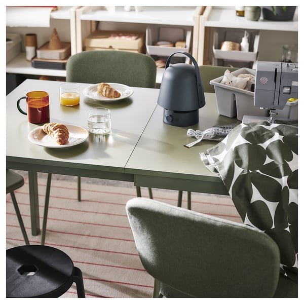 Moderne eetopstelling met groene tafel, roodgestreept vloerkleed, gebak, drankjes, lamp, naaimachine en stof.