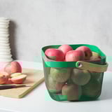 De groene mand van het metaalnetwerk met houten handvat gevuld met rode appels zit op een aanrecht naast een snijplank met gesneden appels en een mes.
