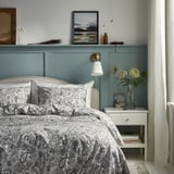Decoratieve slaapkamer met groenblauwe muur, wit beddengoed, paisley dekbed, nachtkastje met bloemen en boeken, en ingelijste kunst.