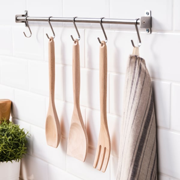 Zes metalen haken hangen houten keukengerei en een gestreepte handdoek aan een wit betegelde muur.