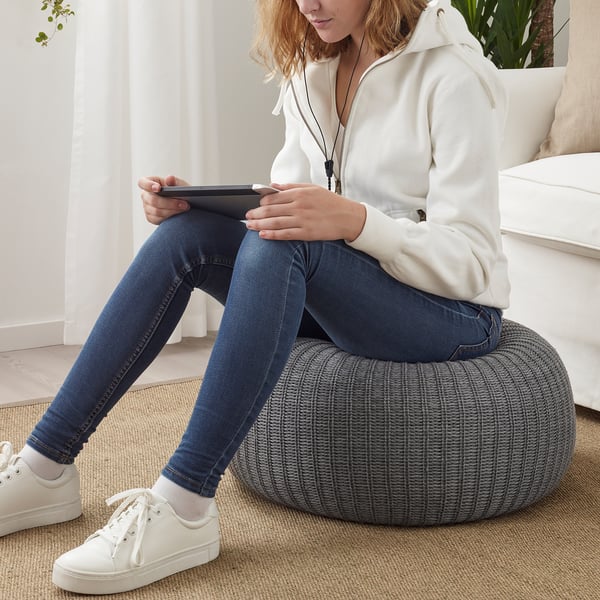 Persoon zittend op ronde voetenbank met behulp van tablet. De persoon draagt een hoodie, een spijkerbroek en sneakers. De kruk is grijs, geweven en dient als extra zitplaats.