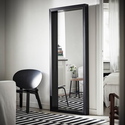 Grote spiegel met zwarte lijst die tegen de muur leunt en een minimalistische kamer weerspiegelt met een zwarte stoel en een gestreept tapijt.