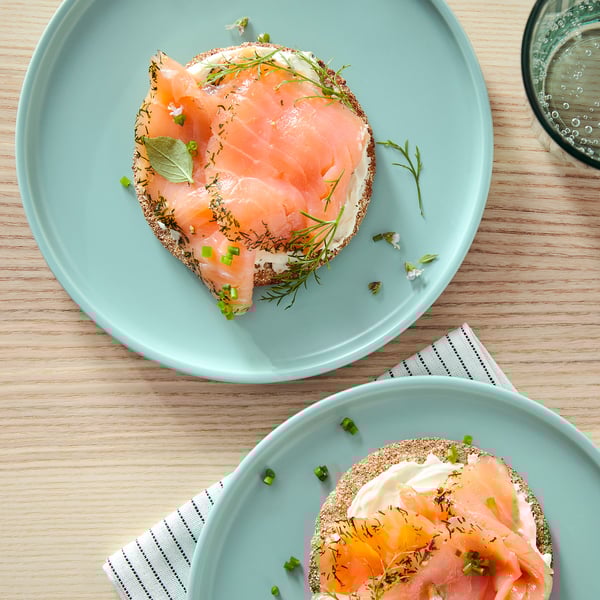 Twee ronde blauwe borden met gerookte zalm, roomkaas, dille en groene uien op crackers, op een houten tafel.