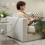 Persoon zittend op de bank, werkend op laptop, omgeven door groen en versierd met kussens. Een lamp en plant op een bijzettafel in de buurt.
