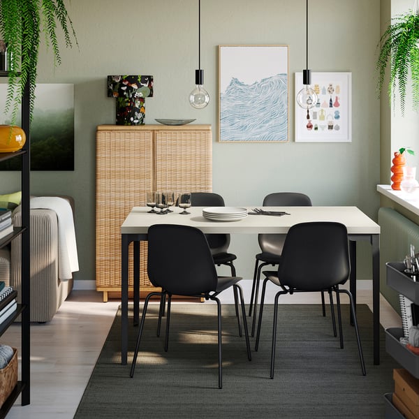 Moderne eetzaal SKÅLSTA witte tafel omringd door zwarte stoelen. Wandgemonteerde rotankast, groen en kleurrijke kunstwerken versterken het interieur.
