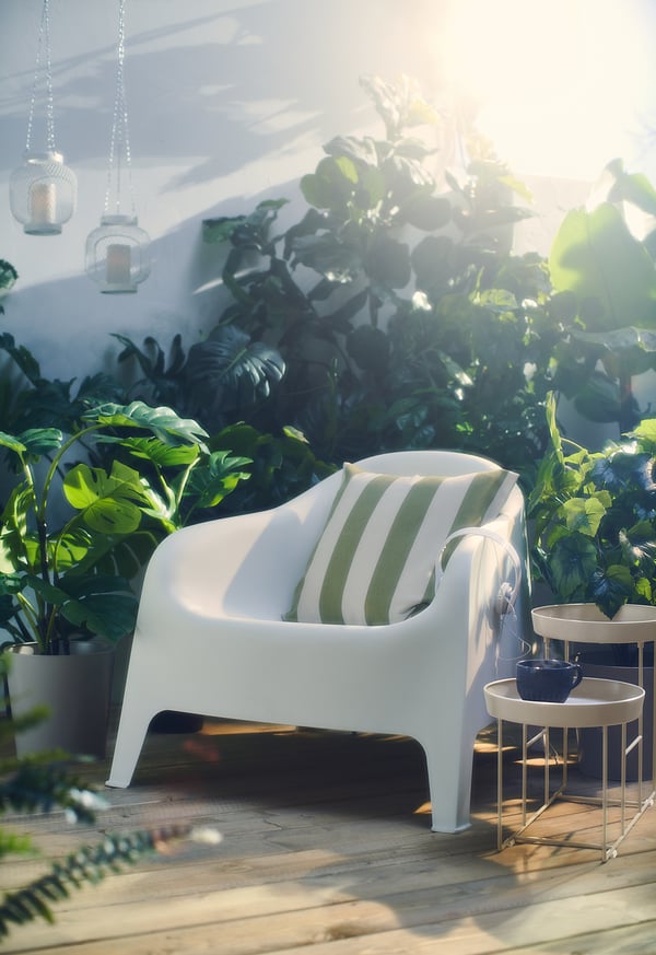 Moderne witte SKARPÖ tuinstoel, gestreept groen kussen, houten terras, weelderig groen, hangende lantaarns.