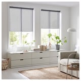 Moderne woonkamer met grote raampartijen, doorschijnende rolgordijnen, witte meubels, planten en een jute vloerkleed.