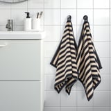Zwart en bruin gestreepte SLÅNHÖSTMAL handdoeken hangen aan haken bij de gootsteen met zeep en tandenborstels.
