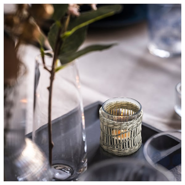 Een SLIPPRIG theelichthouder gemaakt van gevlochten zegge met een brandende kaars, geplaatst naast een heldere glazen vaas met groen op een donkere tafel.