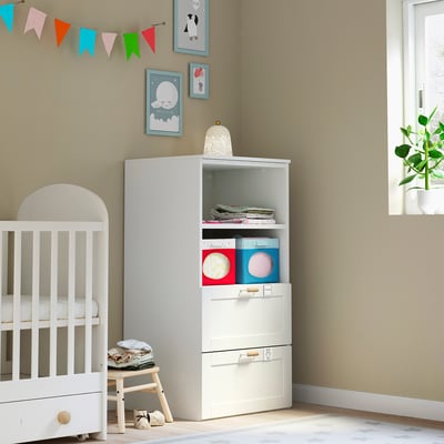 Kleine witte kinderkamer met babybedje, laag krukje en opbergmeubel. Kleurrijk decor en boeken.