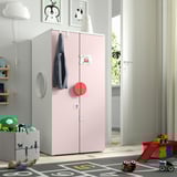 Een lichtroze SMÅSTAD kinderkledingkast met rood handvat, groter dan een speelgoedkist, modern design, zwarte jas, omringd door speelgoed.