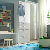 Een kinderkamer met witte SMÅSTAD kledingkast, bureau en lamp, blauw dinosauruskleed, boekenplanken en speelgoedkist.