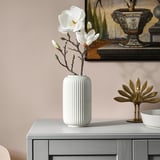Kunstmatige witte magnolia in geribbelde beige vaas op dressoir, met decoratieve gouden pauw; Moderne home decor display.