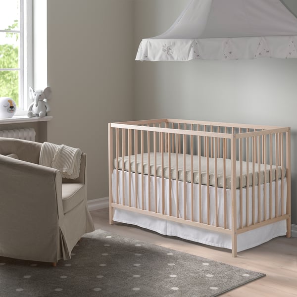 Houten SNIGLAR wieg met verstelbare hoogte in de kinderkamer. Omgeven door grijze fauteuil, gestippeld tapijt, kleine tafel met lamp en speelgoed.