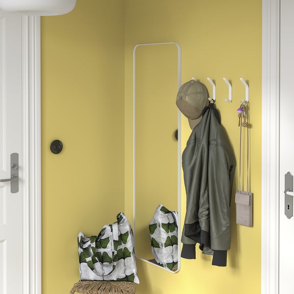 Gele muur met witte SNÖRAKA-haken met groene jas, tas en sleutels. Spiegel onder, rieten bank met kussen.