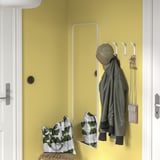 Gele muur met witte SNÖRAKA-haken met groene jas, tas en sleutels. Spiegel onder, rieten bank met kussen.