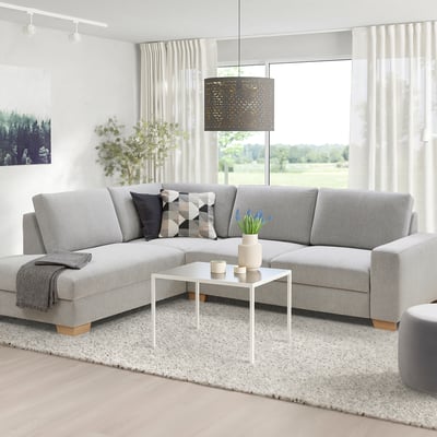 Moderne woonkamer met een grijze SÖRVALLEN sectionele bank, vierkante witte salontafel, kussens met patroon en een groot raam met doorschijnende gordijnen.