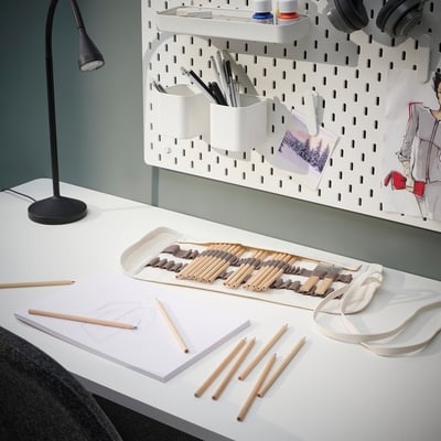 Een wit bureau met SOLFÅGEL kleurpotloden in een houder; een lamp, schetsen en een koptelefoon.