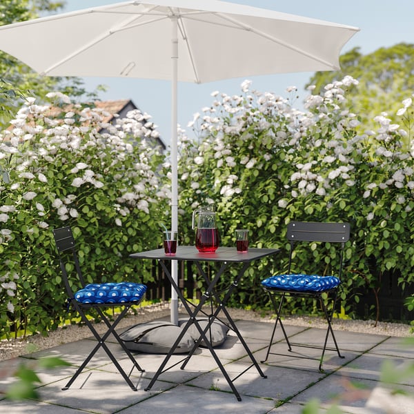 Kleine patioset met klaptafel en stoelen met blauwe kussens, schaduw door witte parasol, tegen een muur van bloeiende witte bloemen.
