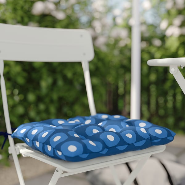 Blauw patroon zitkussen op witte buitenstoel, geplaatst tegen groene gebladerteachtergrond.