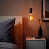 Gezellige slaapkamer met hangende SUNNEBY lamp, warm licht, glazen bol & zwart snoer, naast bed op nachtkastje met boeken & gedroogde bloemen.