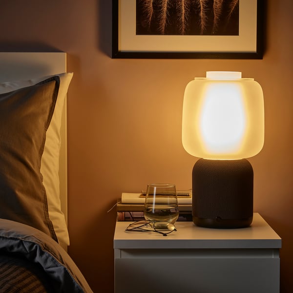 Gezellige slaapkamerhoek met SYMFONISK wifi speakerlamp, bed, boeken, wijn en foto aan de muur.