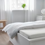 Een netjes opgemaakt bed met witte lakens en twee opgevouwen dekens, tegen een raam met doorschijnende gordijnen. Op de vensterbank staat een kleine potplant.
