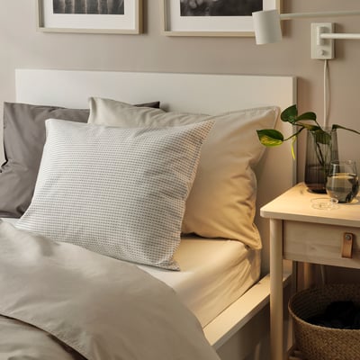 Gezellig bed met witte, grijze en bruine kussens. Nachtkastje met water & plant in aardetinten.