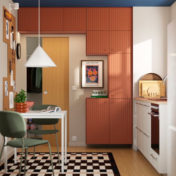 Moderne keuken met TERRSJÖ roodbruine kastzijkanten, witte apparaten, zwart-wit geruit vloer, groene bureaustoel en oranje fruitkunst.