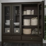 Donkere houten kast met glazen deuren met servies, bestek en stoffen opbergmanden.