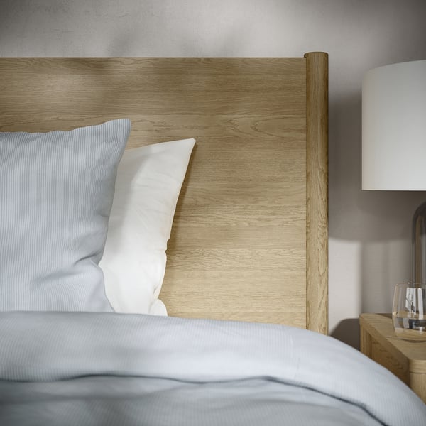 Modern houten bed met strakke lijnen en witte kussens. Op een strak nachtkastje staan een lamp en glas.