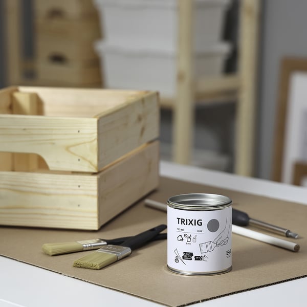 Een doe-het-zelf scène met een klein blikje TRIXIG grijze verf, houten kisten en penselen op een witte tafel.