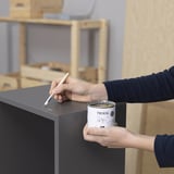Persoon die meubels markeert met pen, met TRIXIG verfblik, in ambachtelijke kamer.