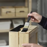 Een persoon brengt zwarte meubelverf aan op een houten oppervlak met behulp van een kwast.