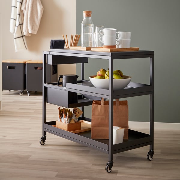 Moderne metalen trolley met drie planken en wielen, met mokken, appels en tassen.