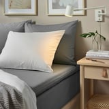 Grijs ULLVIDE bed met katoen & lyocell mix, witte & oranje kussens, bijzettafel met boeken & plant, wandlamp & foto's.