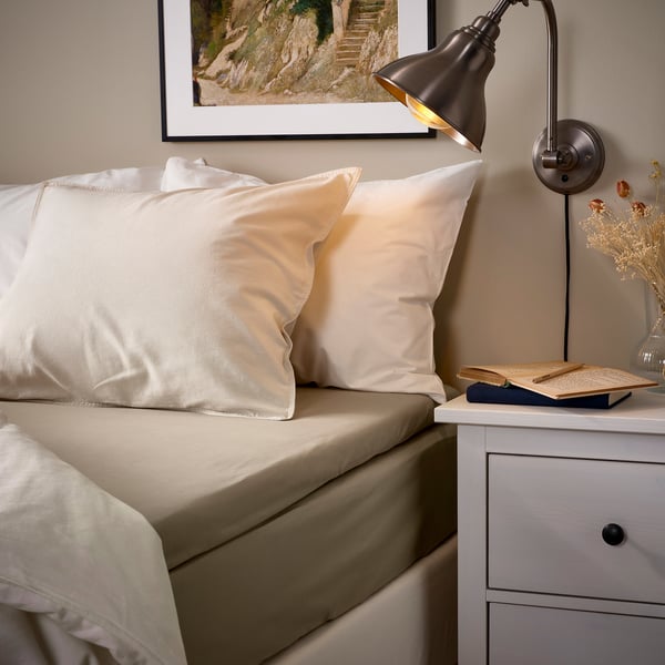 Gezellig bed met beige ULLVIDE beddengoed, wit hoofdeinde, lamp, nachtkastje met boek en bloemen.