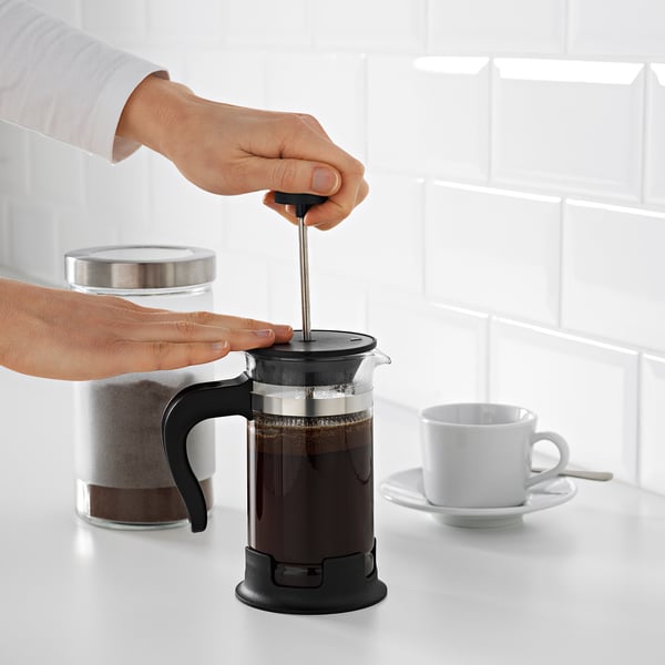 Persoon drukt zuiger op zwart cafetière koffiezetapparaat op witte teller; beker en bus in de buurt.