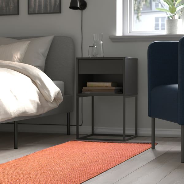 Moderne slaapkamer met een grijs bed, een zwart nachtkastje met boeken en een kleurrijk vloerkleed, dat warmte en textuur aan de kamer toevoegt.