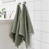 Twee groene VALLASÅN-handdoeken hangen aan muurhaken, langer dan een tandenborstel op een nabijgelegen plank, met een lichtgroene inrichting.