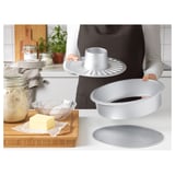 Persoon die bakblikken in keuken houdt. Diverse bakgereedschappen en ingrediënten zichtbaar. Blikken hebben verwisselbare bodems voor verschillende soorten cakes.